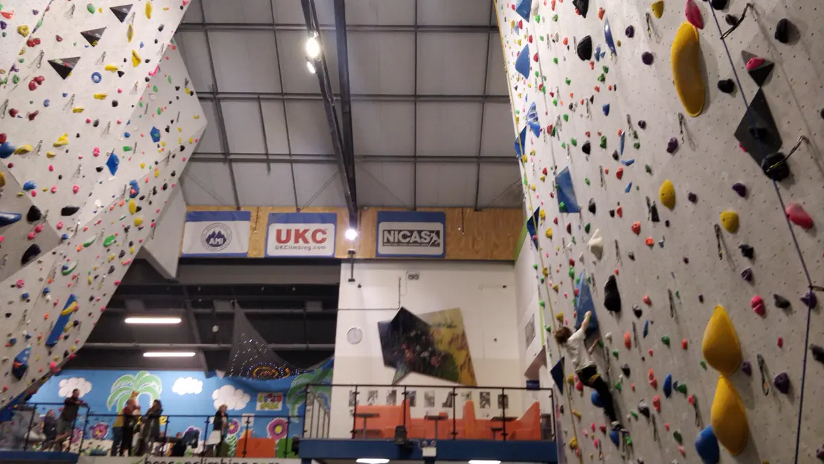 Tall indoor climbing walls covered in holds, with a climber high on the right-hand wall