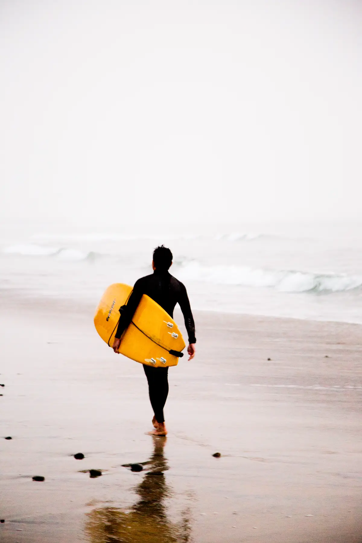 A surfer in a wetsuit carrying a yellow board towards misty waves