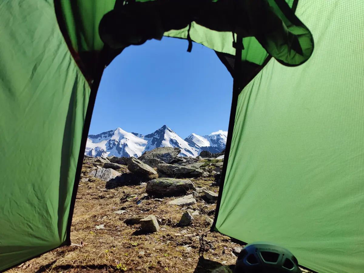 The view through an open tent door to snowy alpine peaks, with a climbing helmet by the entrance