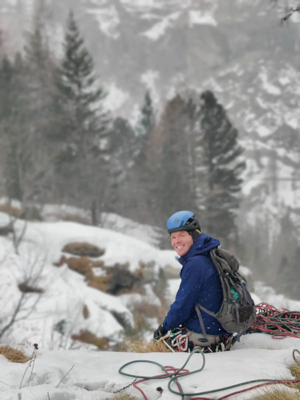 Tom smiling in a blue helmet at a snowy belay, ropes coiled around him, with pine trees behind